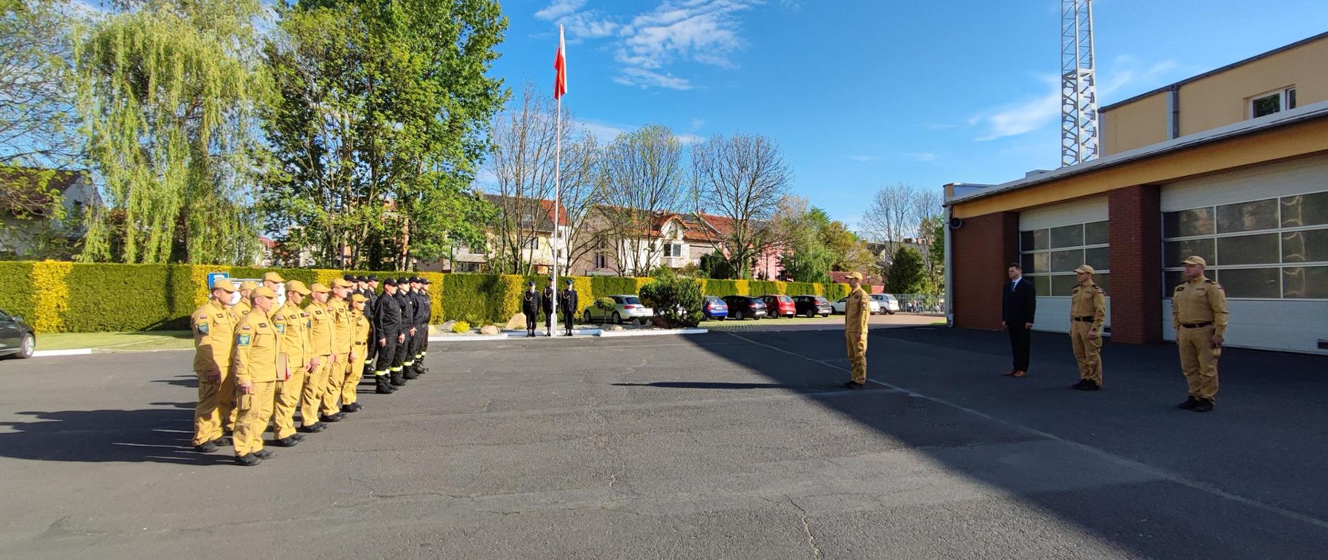 Uczestnicy uroczystości zgromadzeni są na placu Komendy Powiatowej Państwowej Straży Pożarnej w Oleśnie. Na fotografii widoczna również Flaga RP podniesiona na maszt, przy którym ustawiony jest poczet flagowy tworzony przez trzech funkcjonariuszy ubranych w mundur galowy. W tle widoczne budynki i zieleń.