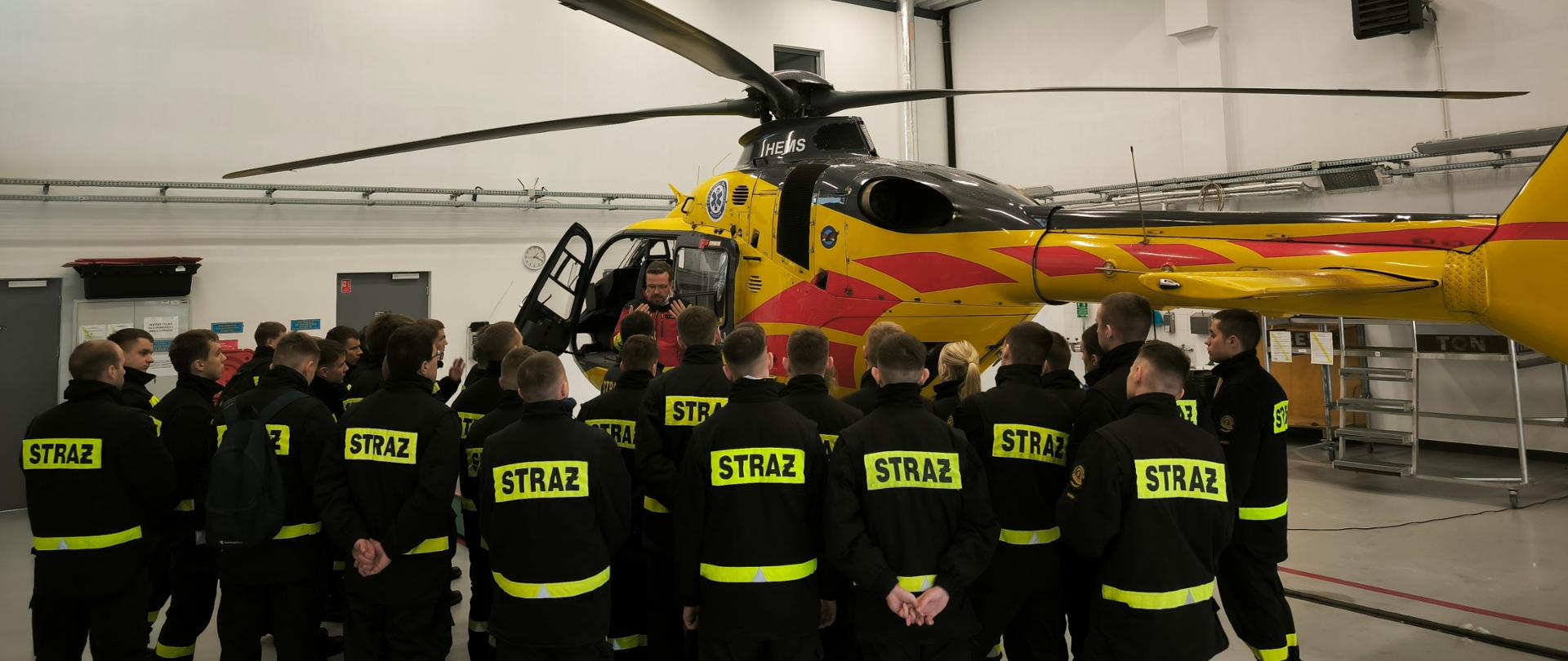 Analiza procedur bezpieczeństwa przy śmigłowcu
Grupa strażaków w czarnych ubraniach koszarowych stoi tyłem, tworząc półkole wokół ogona i kadłuba maszyny. Ratownik LPR prowadzi instruktaż dotyczący bezpiecznych stref poruszania się wokół pracującego śmigłowca.
