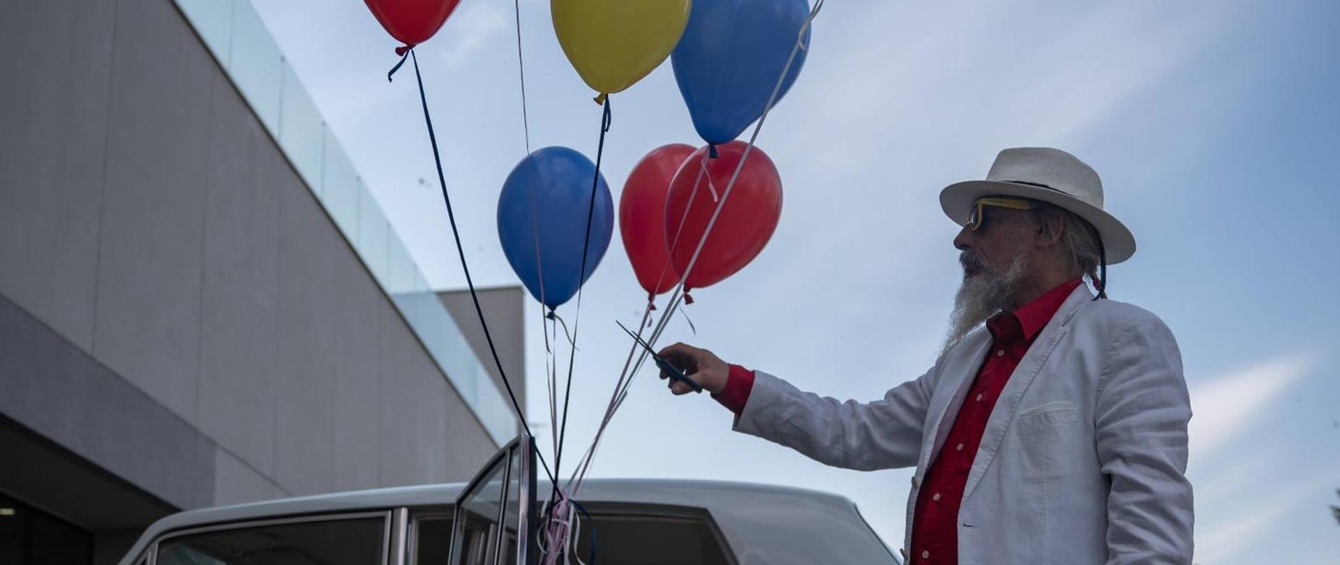 Józef Jan Krużel podczas wystawy „Chromosaturation” przed wejściem do mieleckiej biblioteki w białym garniturze z czerwonym obuwiem, trzymając w ręku pęk kolorowych balonów na tle białego klasycznego samochodu