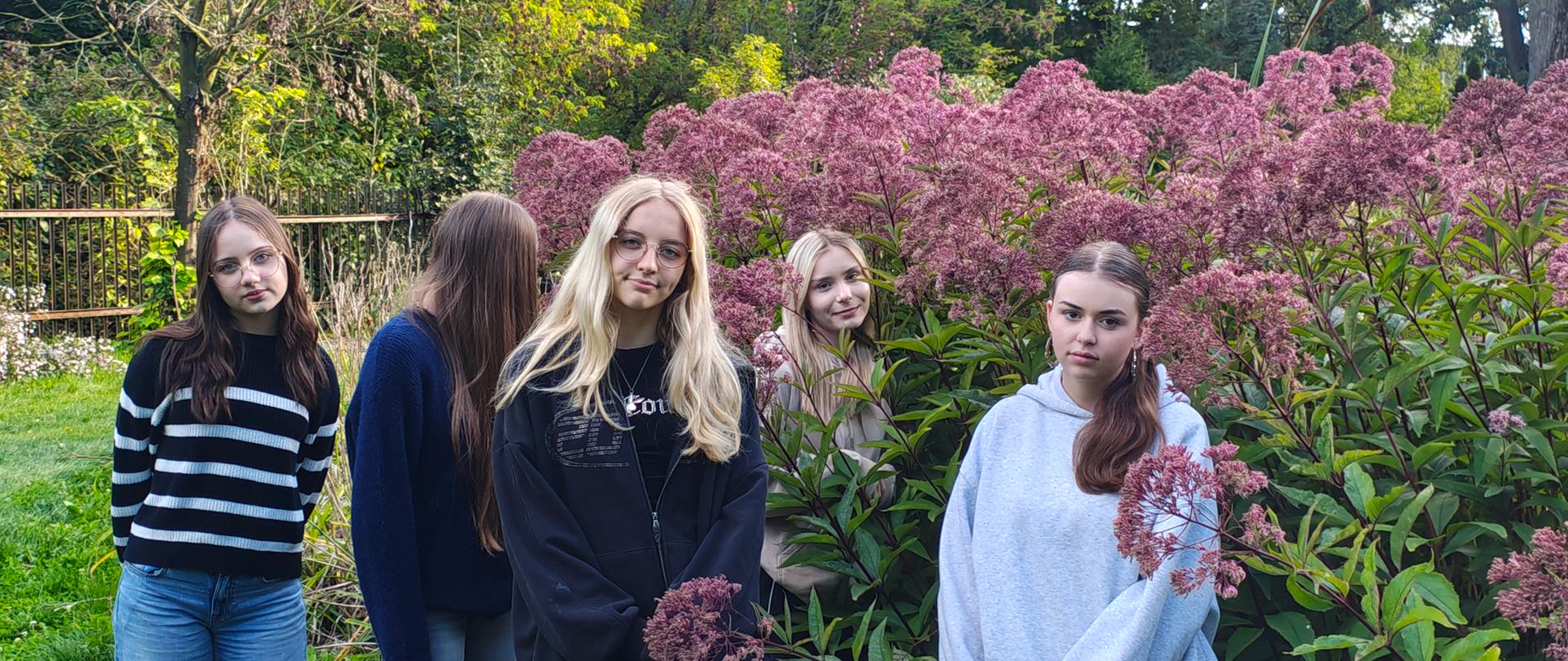 Grupa pięciu dziewczyn stoi wśród różowych i zielonych roślin, cztery dziewczyny patrzą w obiektyw, a jedna jest odwrócona
