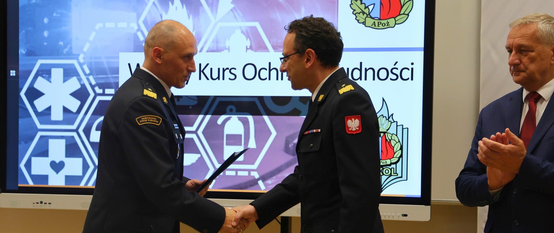 Komendant główny PSP nadbryg. Wojciech Kruczek i rektor-komendant Akademii Pożarniczej nadbryg. dr inż. Tomasz Klimczak wymieniają uścisk dłoni podczas uroczystego zakończenia Wyższego Kursu Ochrony Ludności. W tle ekran z wizualizacją kursu i logotypami WKOL i Akademii Pożarniczej.