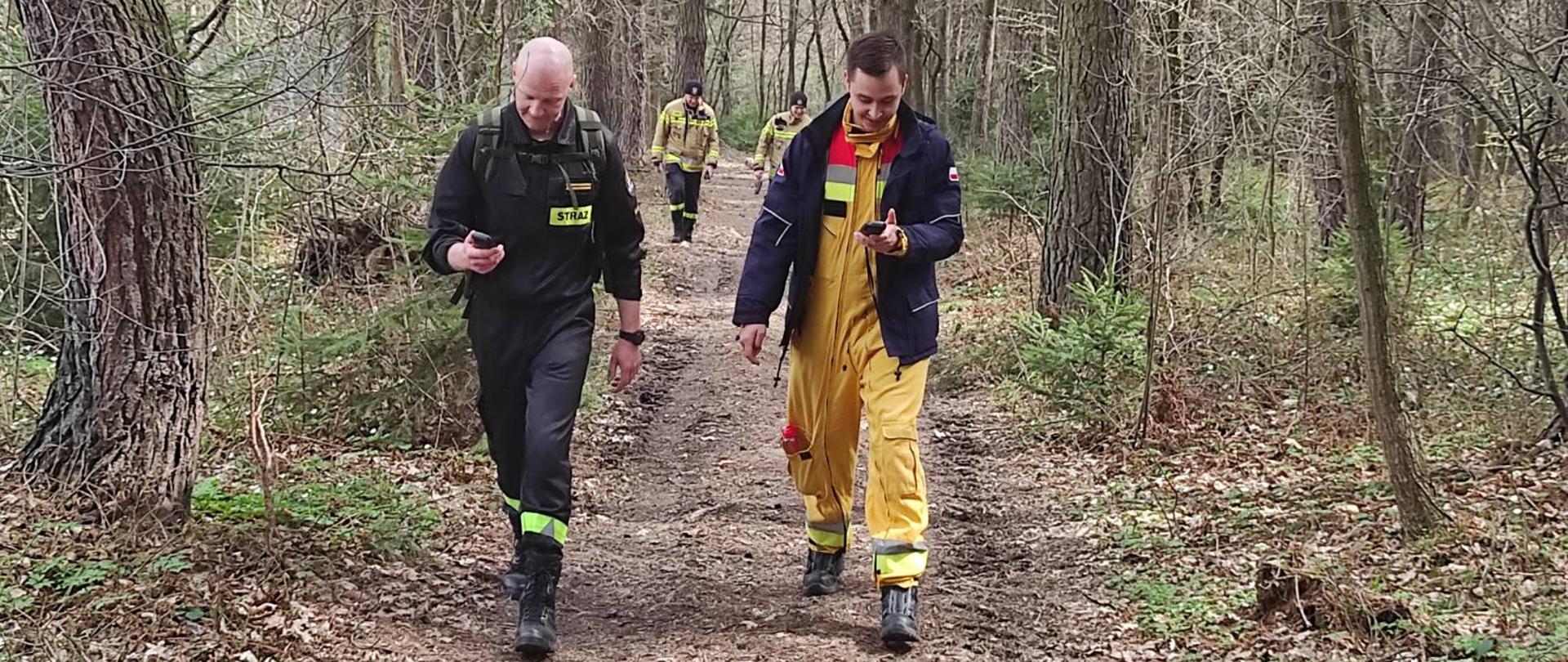 Na zdjęciu widać czterech strażaków, którzy przemieszczają się leśną ścieżką, a w rękach trzymają urządzenia GPS.