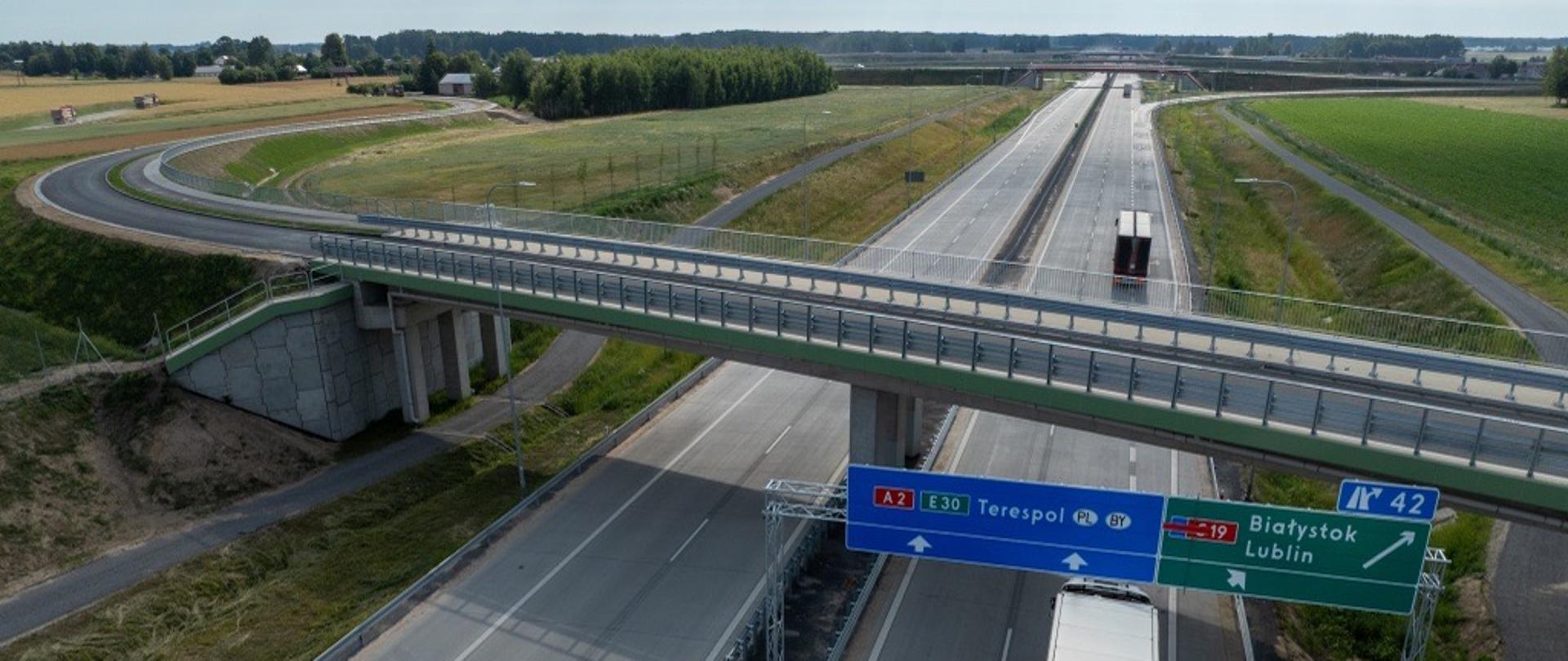 Dwujezdniowa autostrada A2 widziana z góry, z wiaduktem przebiegającym nad jezdniami i ciężarówką jadącą prawym pasem. Nad drogą znajdują się znaki kierunkowe: A2/E30 na Terespol oraz zjazd w kierunku drogi S19 na Białystok i Lublin. Wokół rozciągają się pola i tereny zielone.