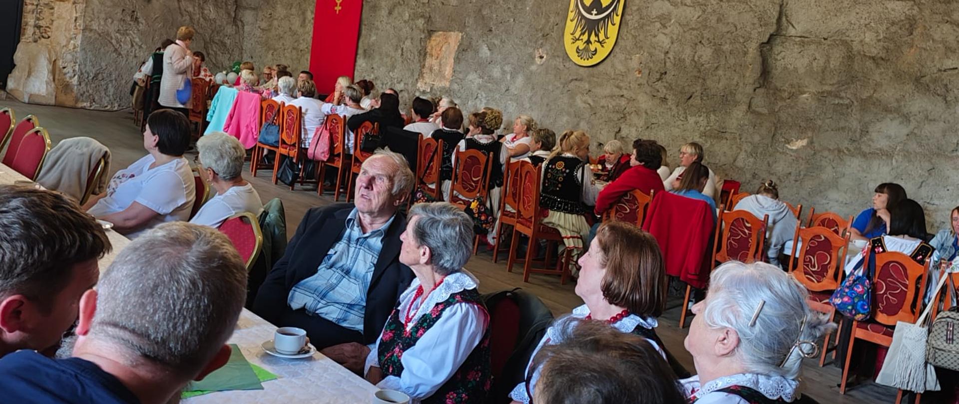 W przestronnej sali o surowych, kamiennych ścianach zgromadzonych jest kilkadziesiąt osób siedzących przy długich stołach. W pierwszym planie widoczne są osoby starsze, część z nich ubrana jest w stroje ludowe. Na ścianach znajdują się czerwony sztandar z dekoracyjnym motywem oraz herb z czarnym orłem na żółtym tle. Pod wysokim sklepieniem wiszą duże, metalowe żyrandole. 