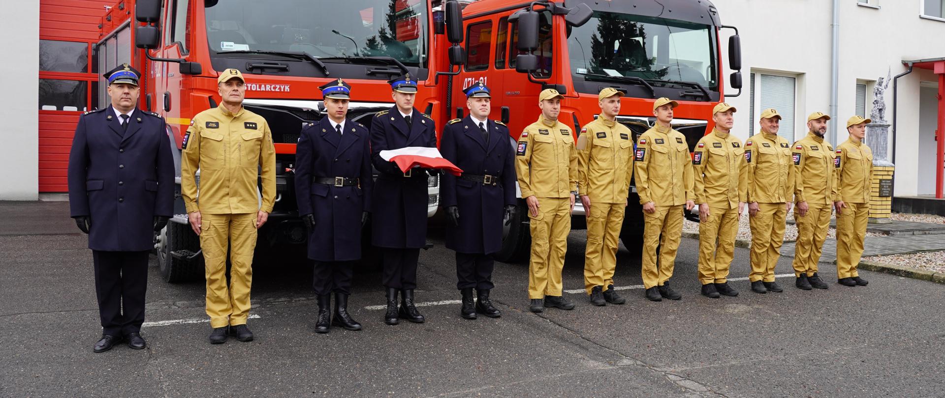 Uroczysta zbiórka przed budynkiem Komendy Powiatowej PSP w Opocznie. Umundurowani strażacy oraz poczet flagowy na tle samochodów pożarniczych. 