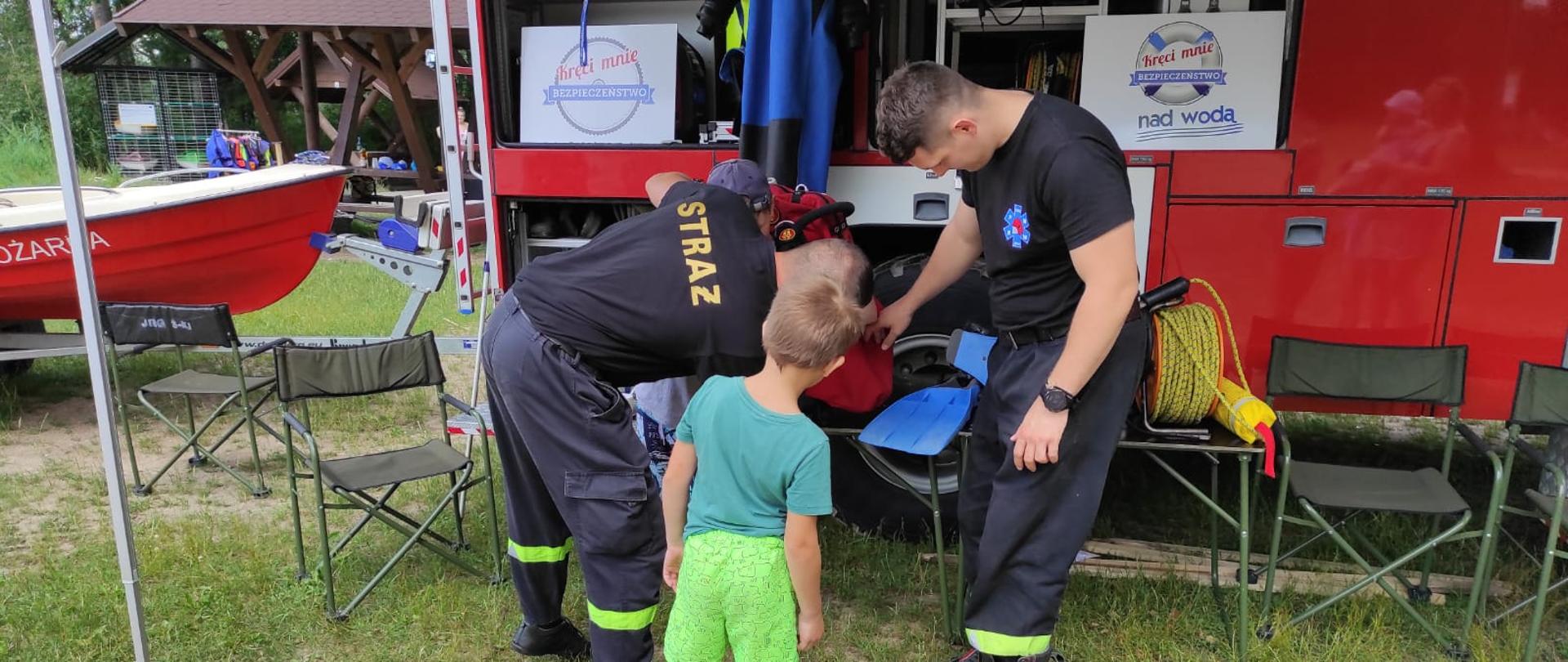 Zdjęcie obrazuje 2 strażaków i dziecko oglądających sprzęt strażacki w tle pojazd strażacki części łodzi i wiaty