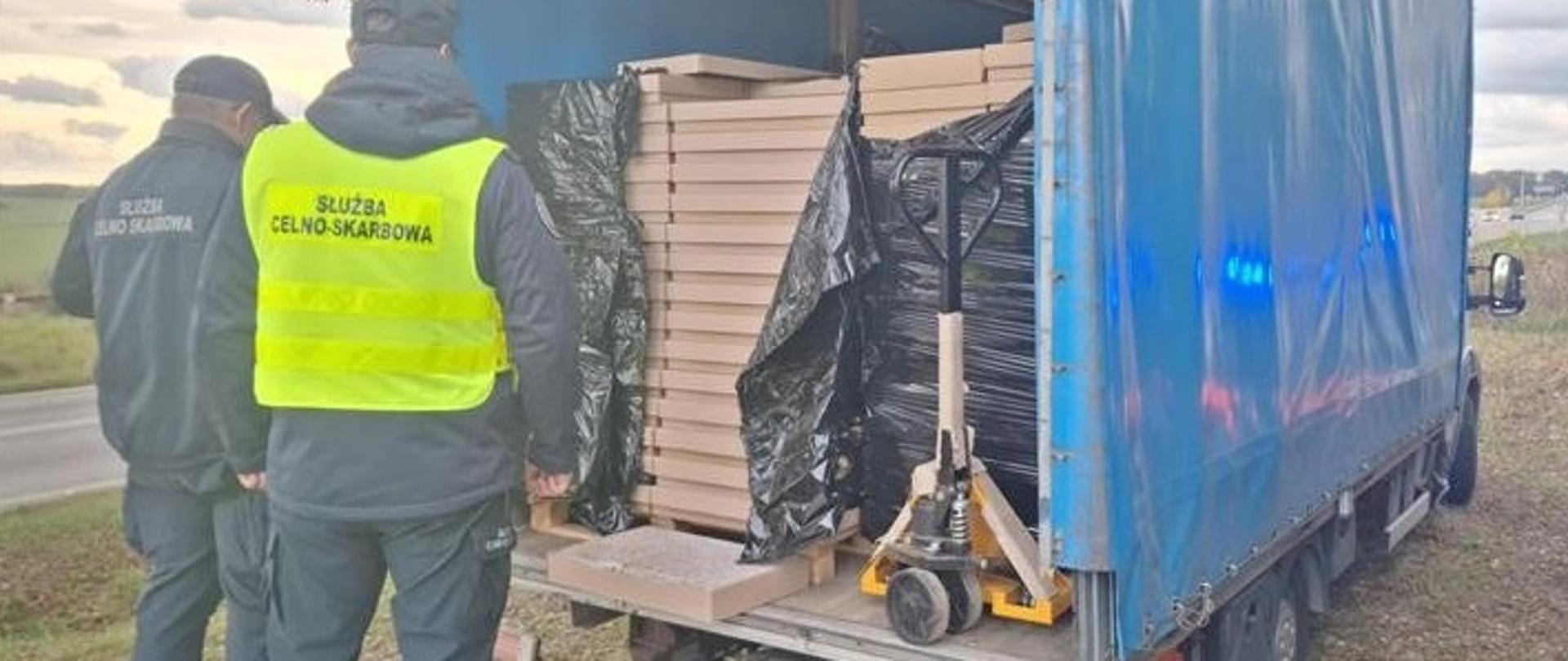 Umundurowani funkcjonariusze stoją przed otwartą przestrzenią ładunkową pojazdu, w którym znajdują się płaskie pudełka kartonowe.