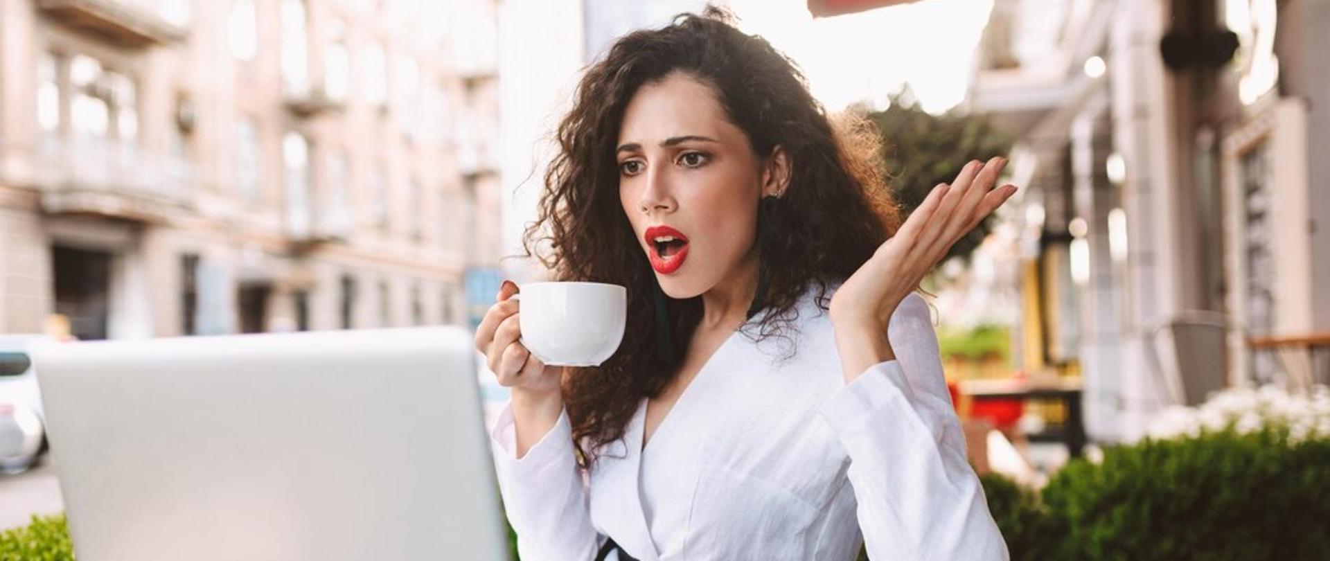 mloda-zszokowana-dama-o-ciemnych-kreconych-wlosach-siedzi-przy-stole-z-filizanka-kawy-w-dloni-i-ze-zdumieniem-patrzy-na-laptopa-w-kawiarni-na-ulicy