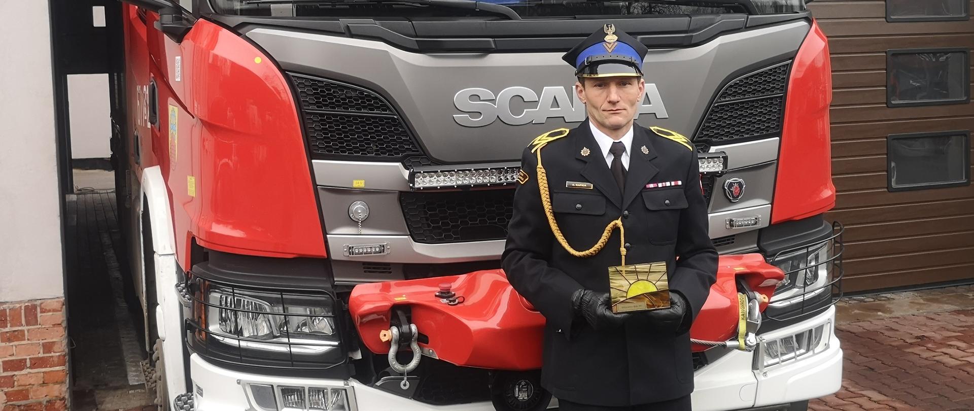 Strażak w pełnym umundurowaniu galowym stoi przed czerwonym wozem strażackim i trzyma w rękach małą, złotą nagrodę w kształcie słońca.