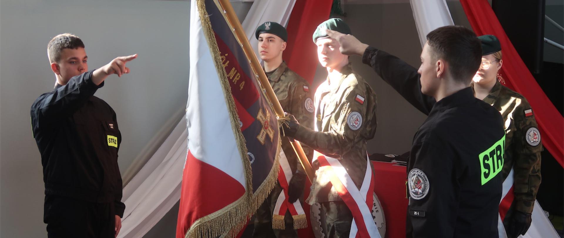 Strażacy składają przysięgę na sztandar szkoły trzymany przez młodzież w mundurach.