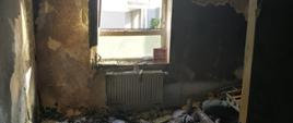 Zdjęcie przedstawia pokój z jednym oknem po pożarze, na podłodze leży popiół, a wszystkie ściany są osmolone. 