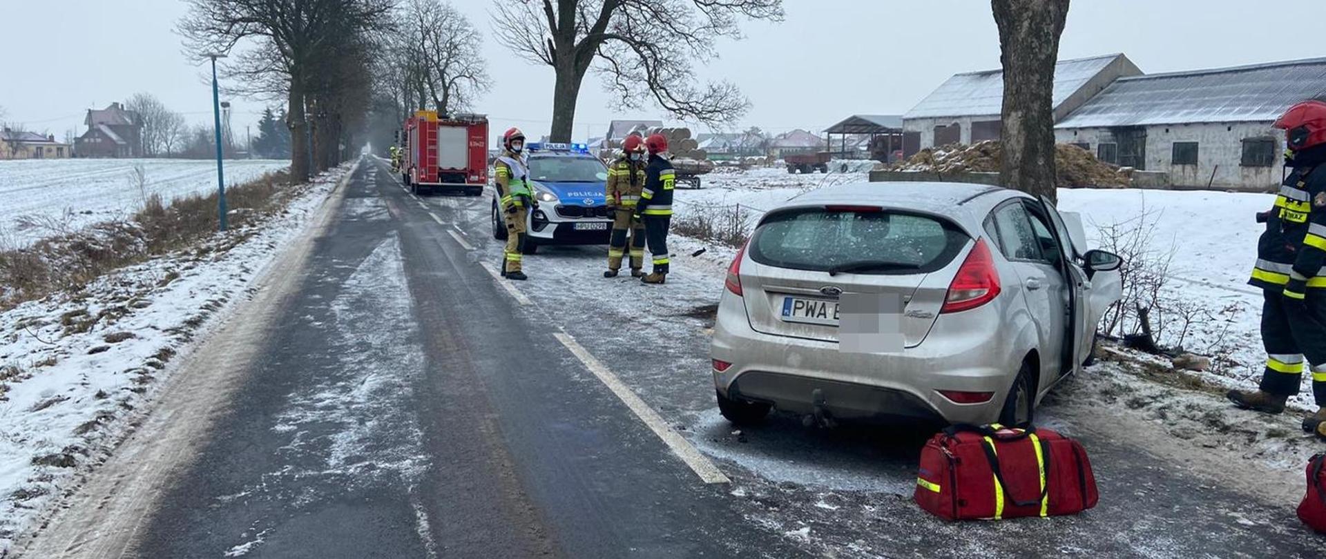 samochód z uyszkodzonym przodem w wyniku wypadku i uderzenia w dzrewo, w tle strażacy i policja