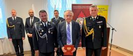 Pomorski komendant wojewódzki Państwowej Straży Pożarnej stoi obok mężczyzny trzymającego czerwoną teczkę i strażaka ochotniczych straży pożarnych za nimi stoją strażacy Państwowej Straży Pożarnej oraz mężczyzna.