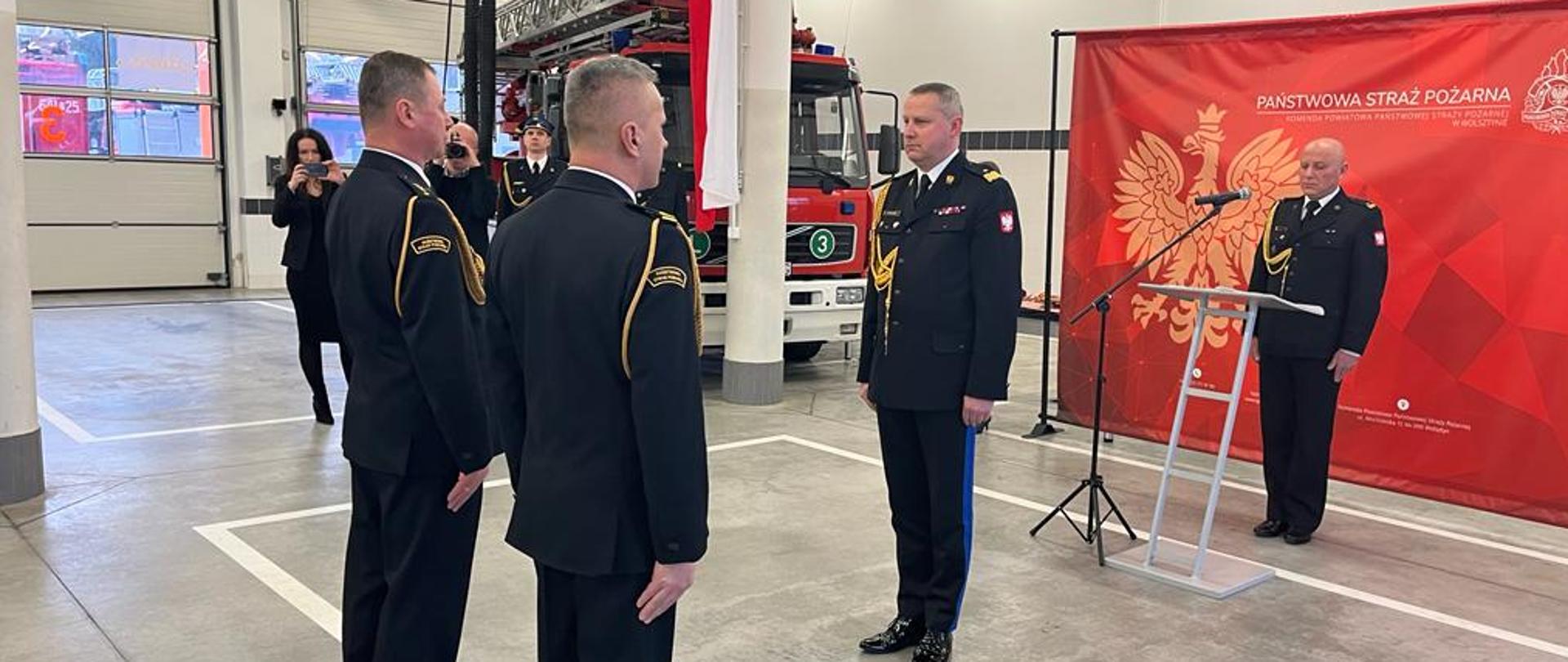 Zdjęcie wykonane podczas uroczystego apelu. Na zdjęciu funkcjonariusze składają meldunek komendantowi wojewódzkiemu PSP. W tle widoczne samochody pożarnicze oraz funkcjonariusze.