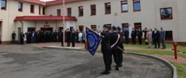 Uroczyste podniesienie flagi państwowej. Poczet flagowy i sztandarowy salutuje. W tle zaproszeni goście przed budynkiem Komendy Powiatowej PSP w Człuchowie.