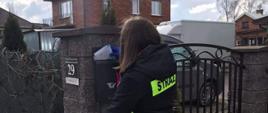 Na zdjęciu widać osobę, druhnę z OSP Stawiszyn, która wkłada ulotkę do skrzynki pocztowej umieszczonej na ogrodzeniu posesji. Kobieta ubrana jest w kurtkę z napisem straż, na rękach ma lateksowe rękawiczki koloru niebieskiego. Na posesji widać pojazdy samochodowe, w dalszej perspektywie budynek z czerwonej cegły. Ochotniczka stoi na kostce brukowej.