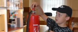 Półkolonia w Śląskowie. W sali wiejskiej odbywają się zajęcia edukacyjne o tematyce związanej z bezpieczeństwem pożarowym. Przed mobilnym symulatorem zagrożeń pożarowych jeden z uczestników prezentuje sposób uzycia gaśnicy proszkowej. Chłopiec trzyma w ręce mikrofon. Na głowie ma czapke dżokejkę koloru czarnego z białym napisem. 