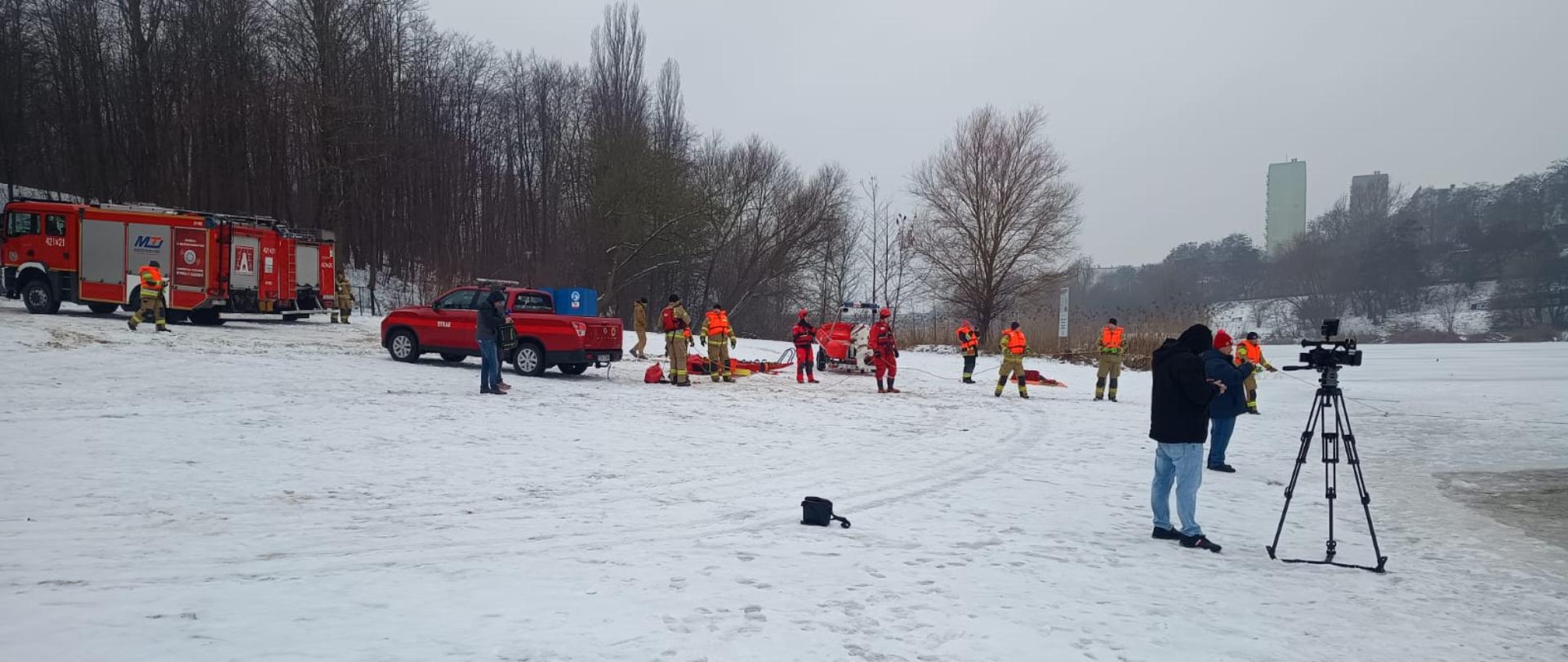 Na zdjęciu widać strażaków oraz sprzęt strażacki, który przygotowywany jest do ćwiczeń. Widać również operatora TV oraz przedstawicieli mediów, którzy przybyli na zaproszenie tutejszej komendy.