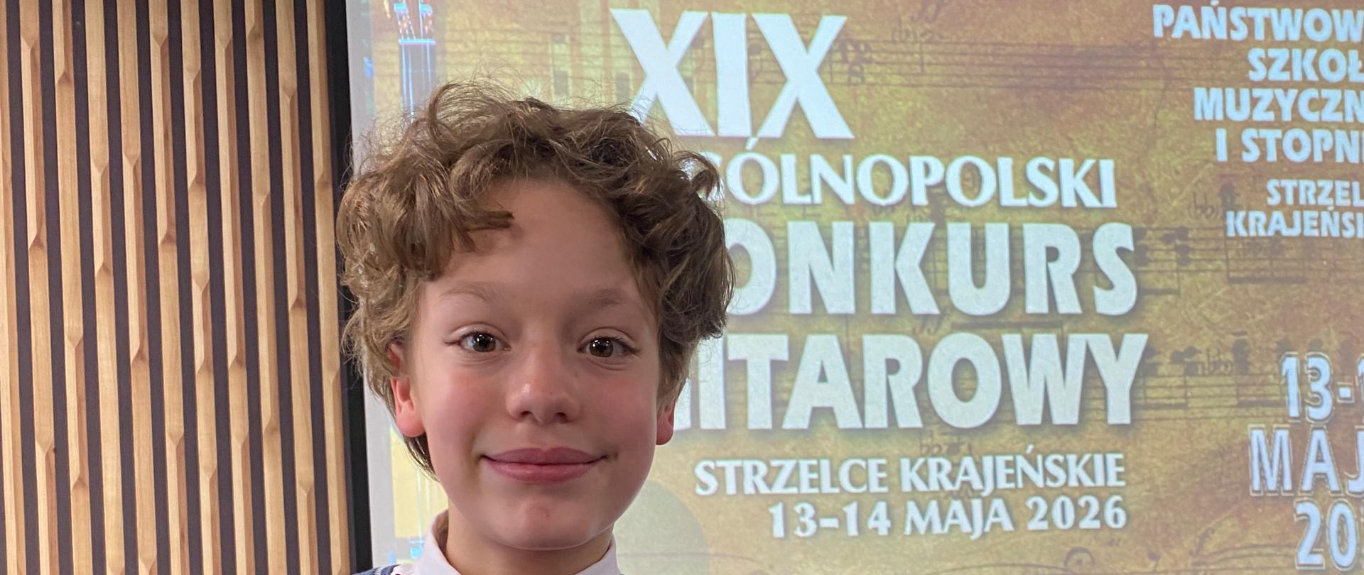 Zdjęcie przedstawia ucznia z dyplomem i futerałem na tle plakatu XIX Ogólnopolskiego Konkursu Gitarowego w Strzelcach Krajeńskich, który odbył się w dniach 13-14 maja 2026 r.