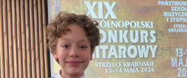 Zdjęcie przedstawia ucznia na tle plakatu XIX Ogólnopolskiego Konkursu Gitarowego w Strzelcach Krajeńskich, który odbył się w dniach 13-14 maja 2026 r.