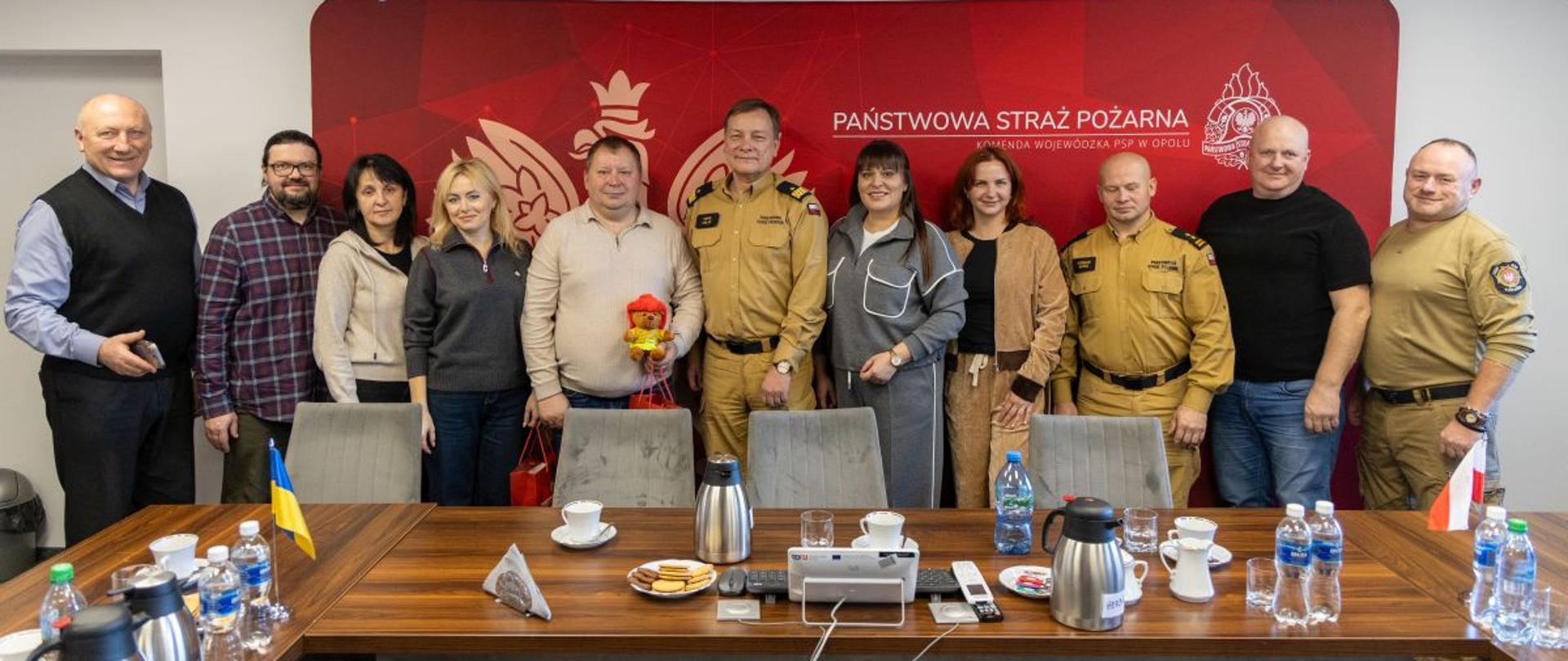Grupa jedenastu osób, w tym funkcjonariusze PSP i ukraińscy samorządowcy, pozuje do wspólnego zdjęcia w sali konferencyjnej. W tle widoczny jest napis „PAŃSTWOWA STRAŻ POŻARNA”. Na stole znajdują się flagi Polski i Ukrainy oraz napoje.