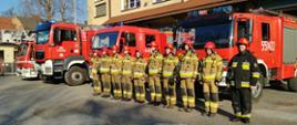 Widok z przodu. Dziesięciu strażaków JRG Wadowice stoi w ubraniach specjalnych przed samochodami pożarniczymi przed garażami JRG. 