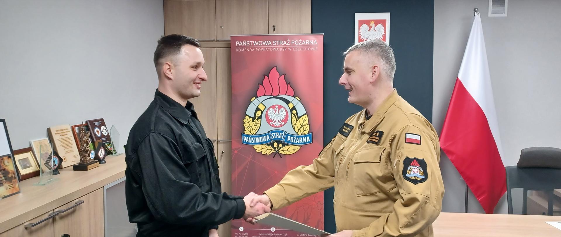 Komendant Powiatowy Państwowej Straży Pożarnej w Człuchowie st. bryg. Radosław Batruch stoi w gabinecie i wręcza akt mianowania na stanowisko ratownika w służbie stałej st. str. Dawidowi Synoradzkiemu.