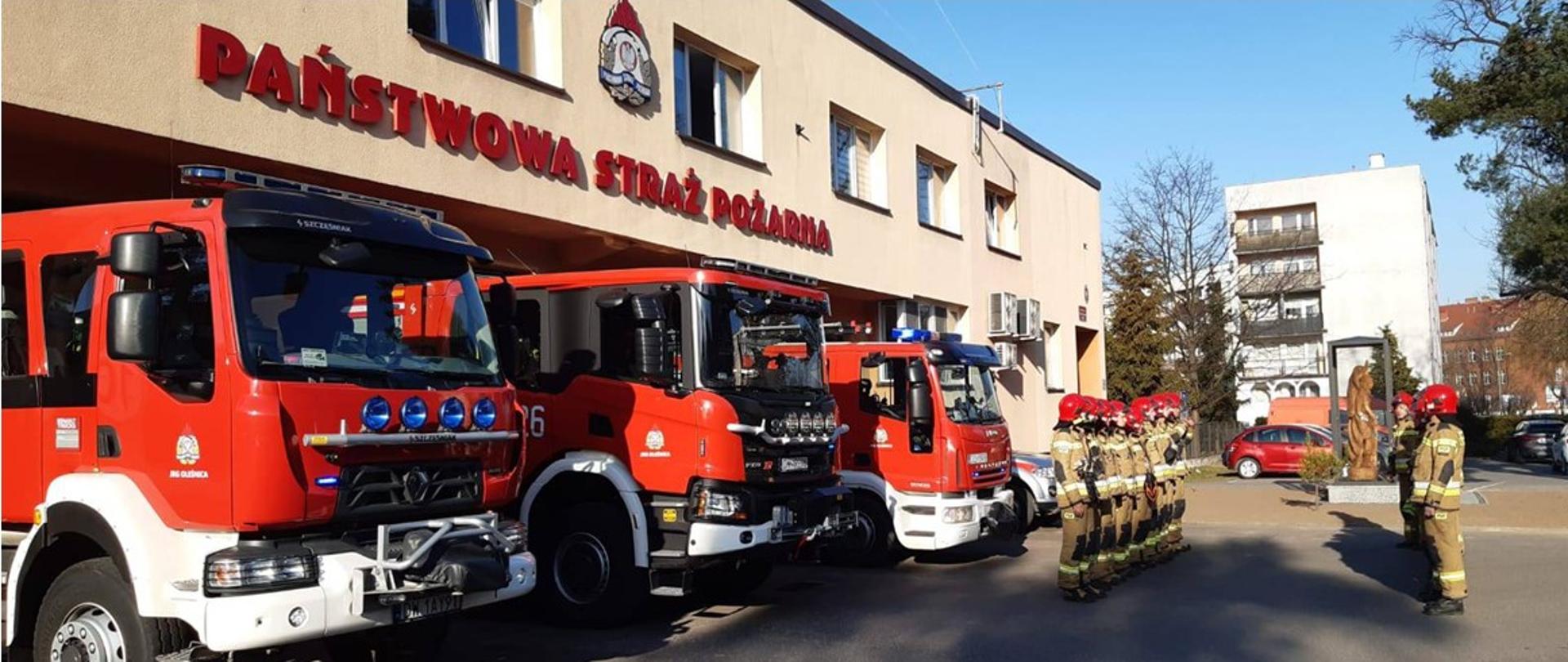Na zdjęciu widać samochody strażackie oraz strażaków stojących w szeregu na baczność w czasie oddawania hołdu poległym strażakom z Ukrainy. w tle widać figurę świętego Floriana. 