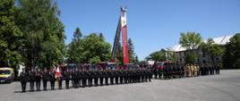 Pododdziały strażaków stoją na placu wewnętrznym Szkoły podczas uroczystości, w głębi stojące wozy strażackie, z samochodu typy drabina zwisa baner z logo 30-lecia CS PSP, logo CS PSP oraz napisem Centralna Szkoła Państwowej Straży Pożarnej