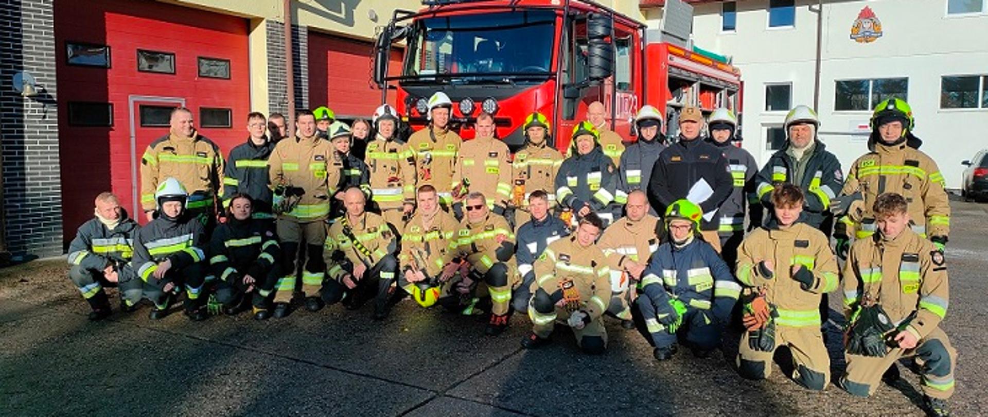 Grupa strażaków ubrana w ubrania specjalne w kolorze piaskowym oraz granatowym pozuje do zdjęcia na tle samochodu ratowniczo-gaśniczego. Scena dzieje się na placu manewrowym przy budynku o elewacji w kolorze żółtym. W elewacji znajduje się szereg dużych czerwonych bram garażowych. Widać błękitne, bezchmurne niebo.