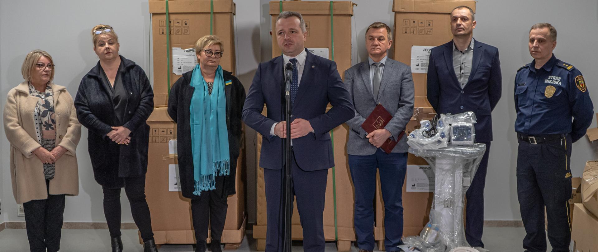 Jeden mężczyzna stojący przed mikrofonem i wypowiadający się. Za nim trzy kobiety i trzech mężczyzn