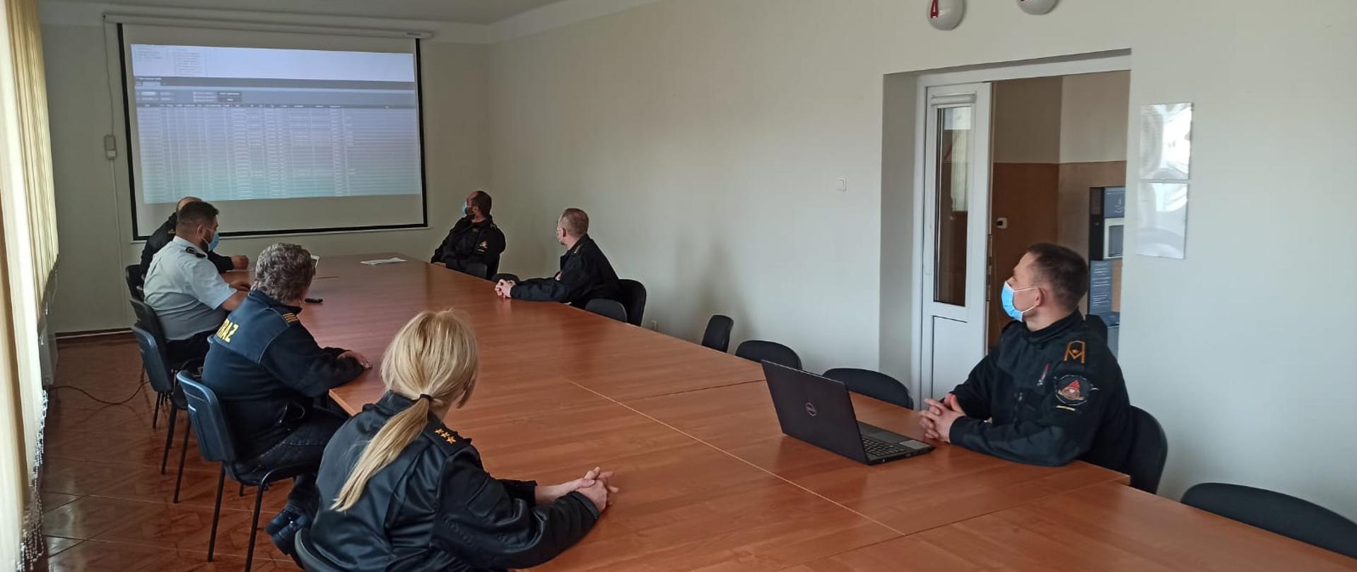Zdjęcie przedstawia szkolenie, które odbywa się w sali szkoleniowej. W centrum zdjęcia widać stoły przy których siedzą funkcjonariusze uczestniczący w szkoleniu ubrani w czarne mundury oraz maseczki chirurgiczne. Jena osoba prowadząca szkolenie, która siedzi przy komputerze jest ubrana w niebieską służbową koszulę. W tle zdjęcia widać: drzwi, lampy alarmowe, rzutnik, tablice projekcyjną na której wyświetlony jest program do łączności, żaluzje okienne.