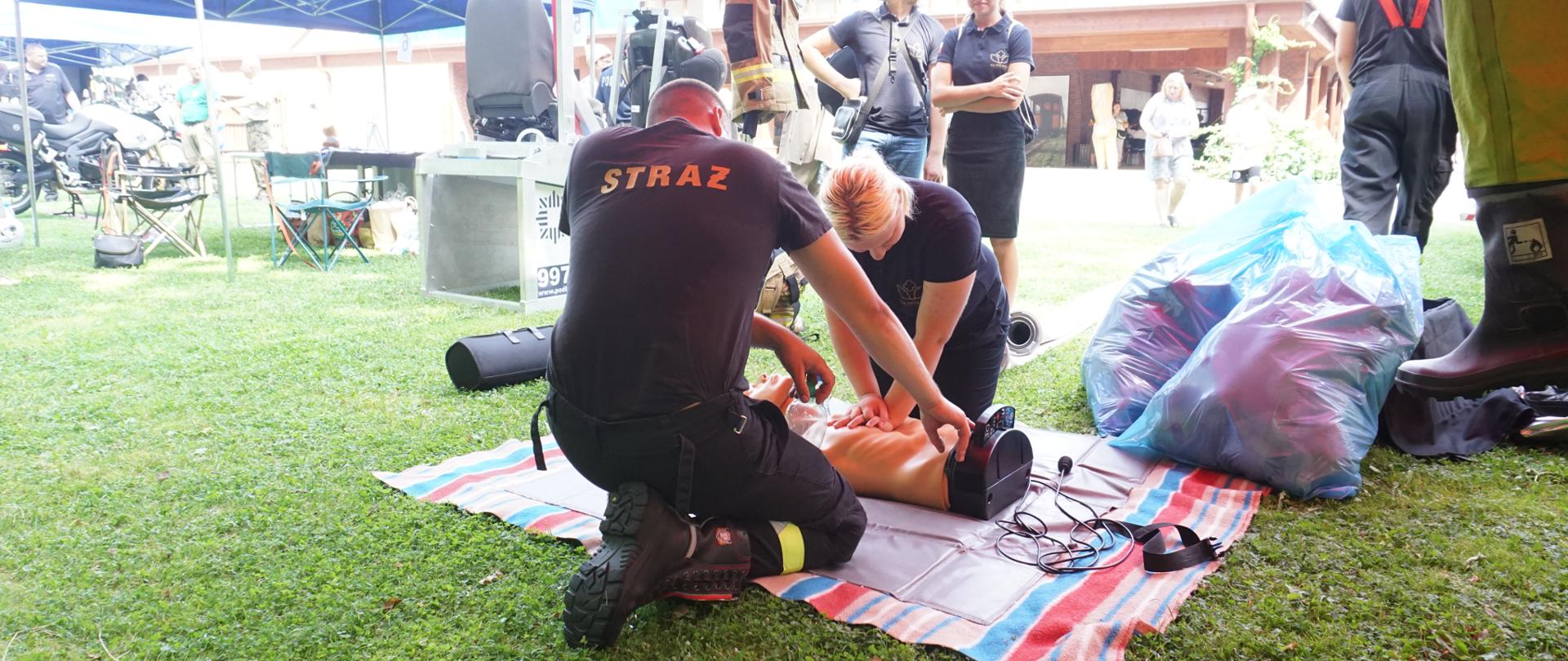 W pierwszym tle strażak udziela instruktażu wykonywania zewnętrznego masarzu serca na fantomie.
