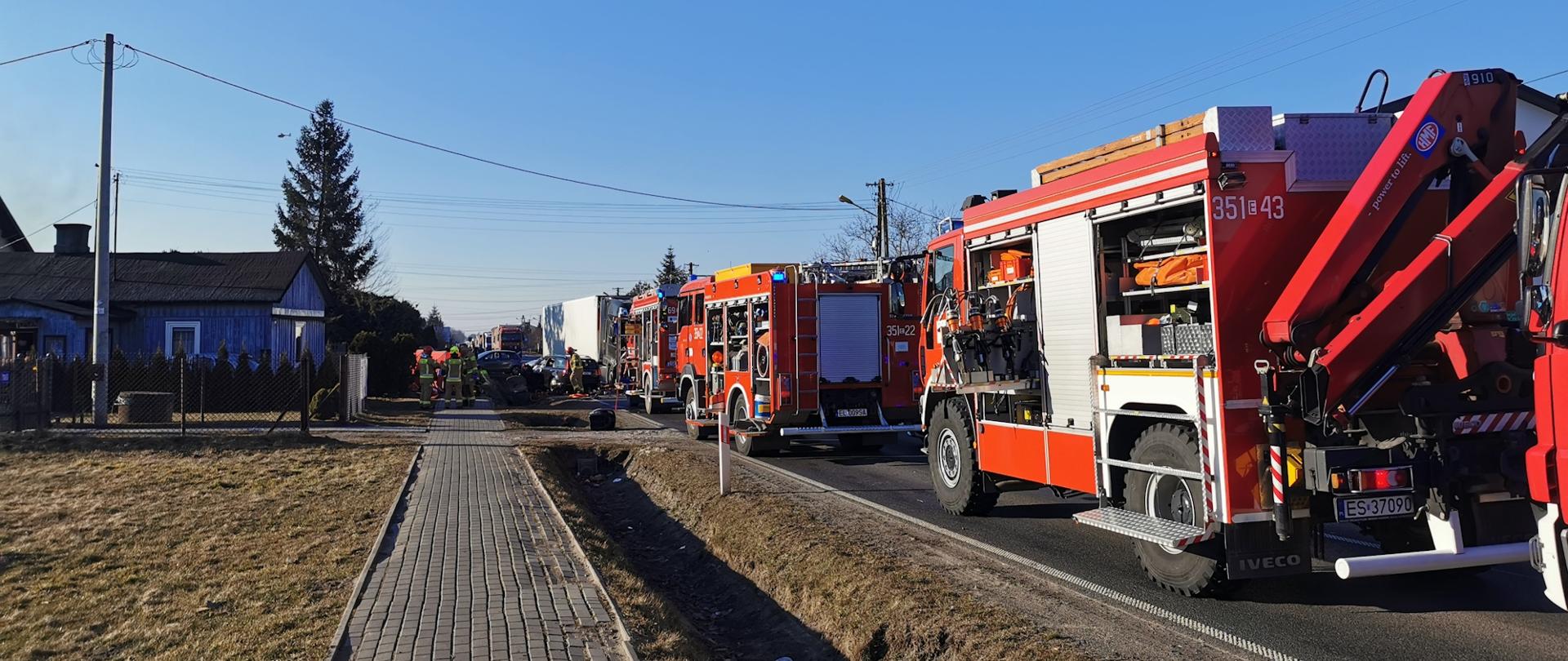Na zdjęciu cztery pojazdy strażackie koloru czerwonego, w tle widać budynek mieszkalny, działania ratownicze i rozbite pojazdy. 