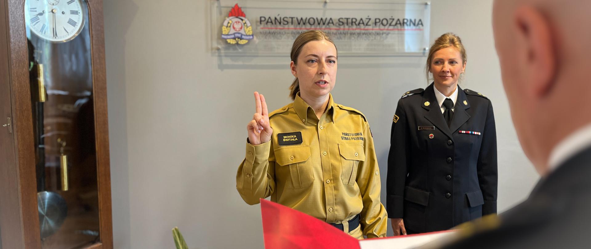 Kobieta w piaskowym mundurze Państwowej Straży Pożarnej składa ślubowanie, unosząc dwa palce prawej dłoni w górę. Za nią stoi uśmiechnięta kobieta w ciemnogranatowym mundurze wyjściowym, a na ścianie widnieje tablica z napisem „Państwowa Straż Pożarna”. Na pierwszym planie po prawej stronie widać tył głowy i ramię mężczyzny w mundurze, który trzyma otwartą, czerwoną teczkę. W tle po lewej stoi wysoki, stojący zegar szafowy.