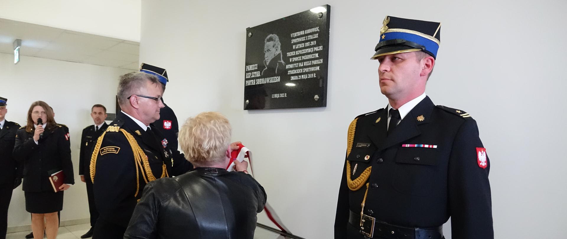 komendant i wdowa po strażaku zdejmują wstęgę z tablicy. Tablica ma asystę oficerów PSP.