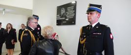 Komendant i wdowa po strażaku zdejmują wstęgę z tablicy. Tablica ma asystę oficerów PSP.