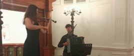 A performance of two artists: Karolina Mikołajczyk stands on the left in a long black dress, playing the violin; Iwo Jedencki sits on the right, playing the accordion.
