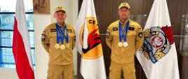 Dwóch strażaków Centralnej Szkoły PSP-uczestników zawodów European Police & Fire Games stoi obok siebie ze zdobytymi medalami na szyi, pomiędzy nimi od lewej strony flaga Polski, flaga z logo CS PSP, flaga z logo PSP