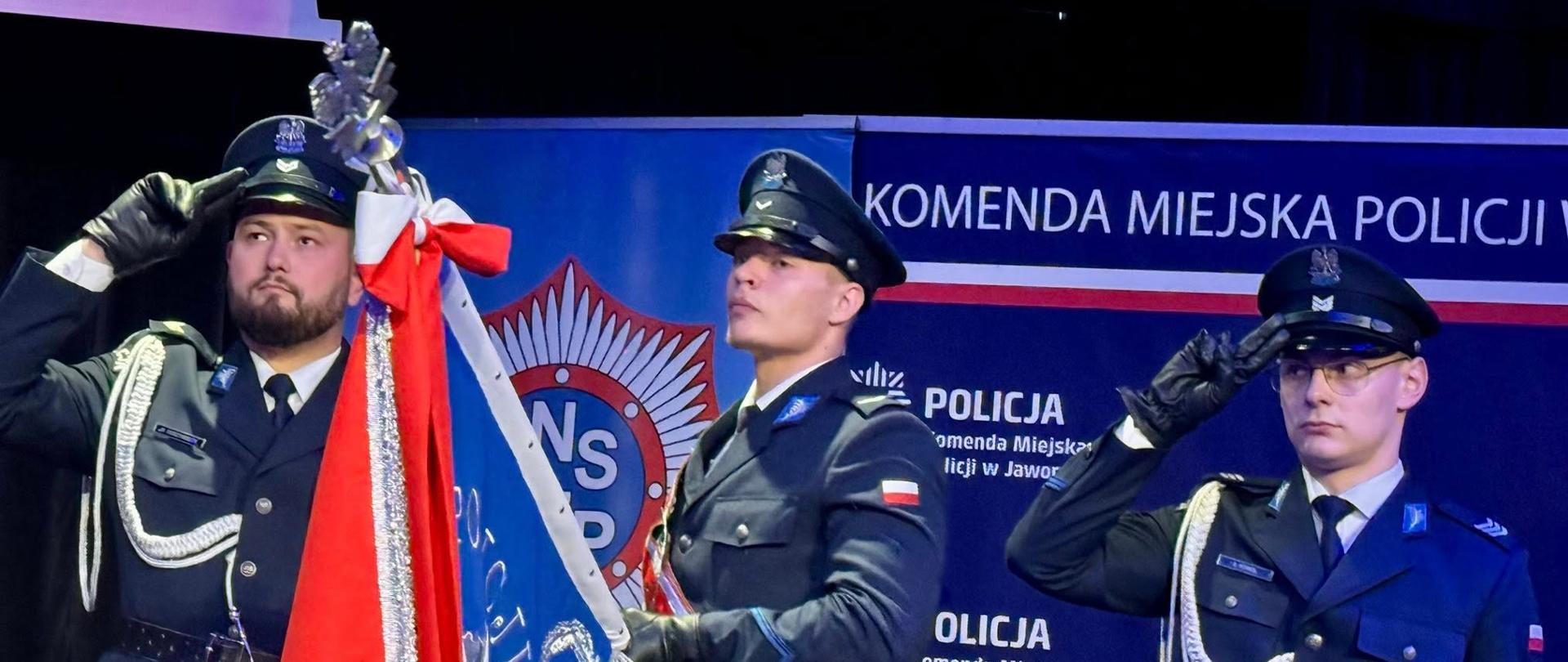 Trzech policjantów w galowych mundurach stoi na baczność przy policyjnym sztandarze podczas uroczystości.
