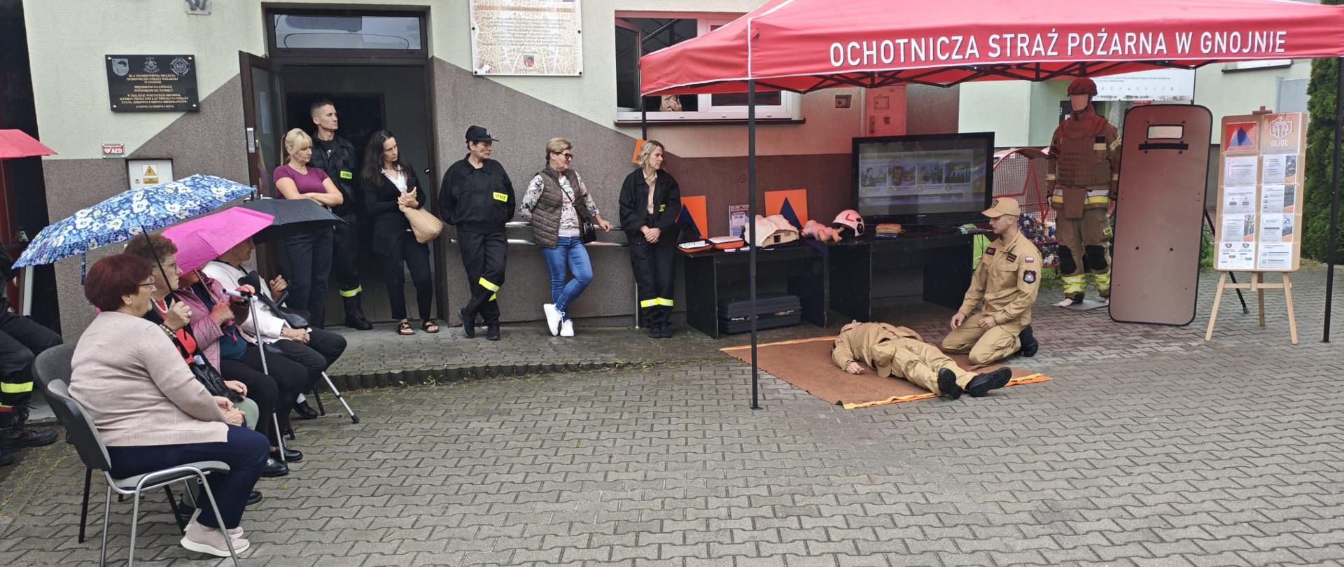 Szeroki plan sprzed remizy OSP w Gnojnie. Przed budynkiem ustawiono czerwony namiot edukacyjny, gdzie prowadzony jest pokaz pierwszej pomocy z udziałem instruktora i leżącego manekina. Po lewej stronie na krzesłach siedzą widzowie pod parasolami, a pod ścianą budynku stoi grupa obserwujących osób.