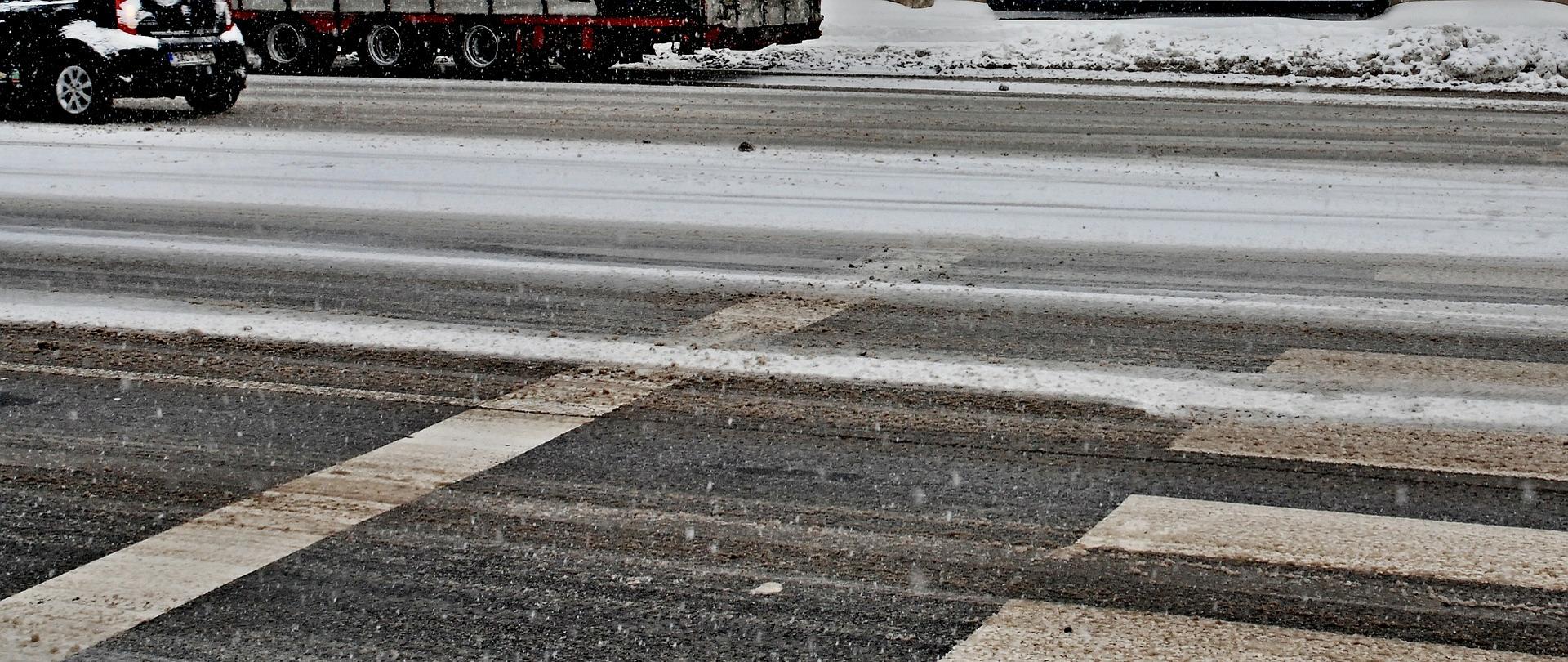 Na zdjęciu po prawej stronie przejście dla pieszych. Widać ośnieżoną drogę. Po lewej stronie fragmenty samochodów. 