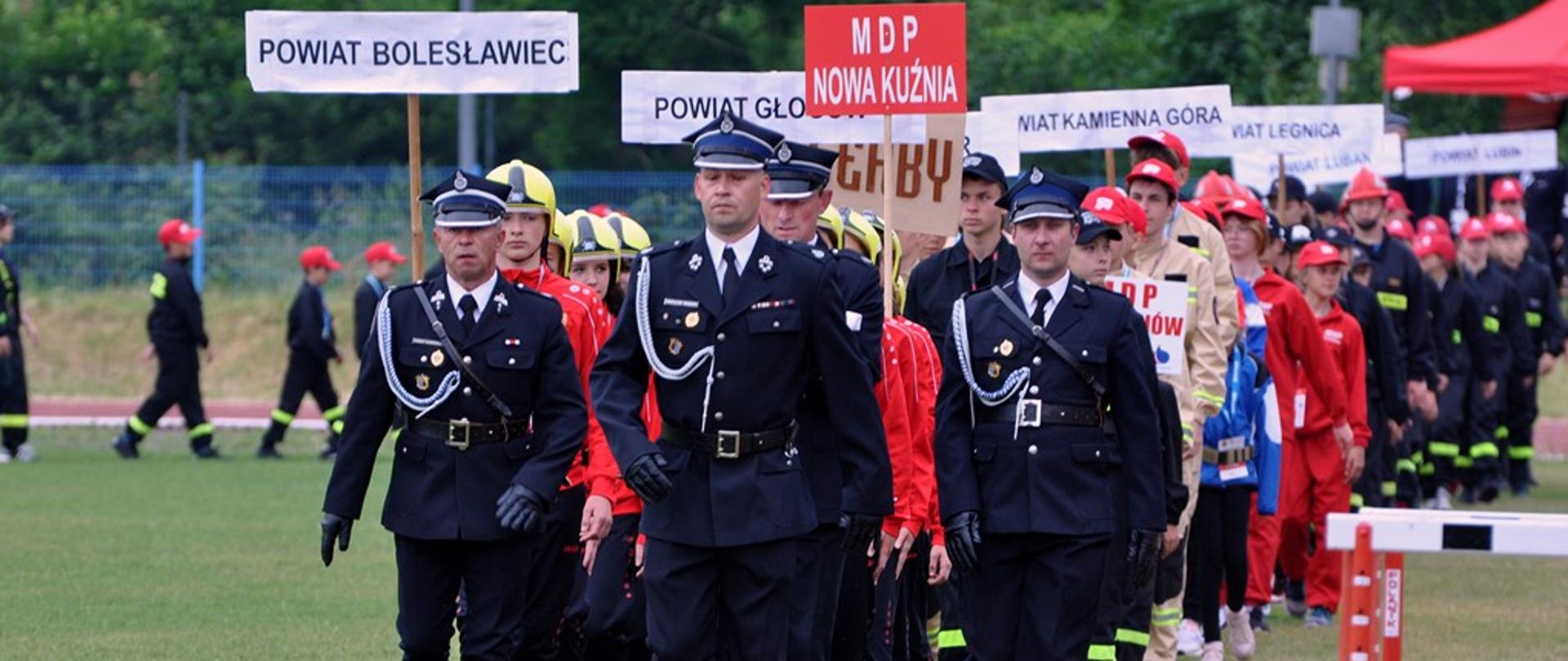 Dzień. Widok na murawę boiska. Po której maszerują w dwuszeregu grupa dzieci. Nad dziećmi widać tabliczki z nazwami powiatów. Na przedzie grupy idzie druh ubrany w umundurowanie galowe, srebrny sznur galowy, rogatywkę oraz czarne, wysokie buty. Za nim idzie w szeregu trzech druhów.