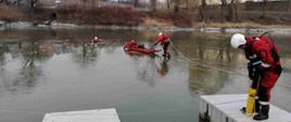zdjęcie przedstawia zamarznięty zbiornik wodny na którym leży jeden strażak i woła o pomoc. Drugi strażak z brzegu rzuca mu rzutkę. 
