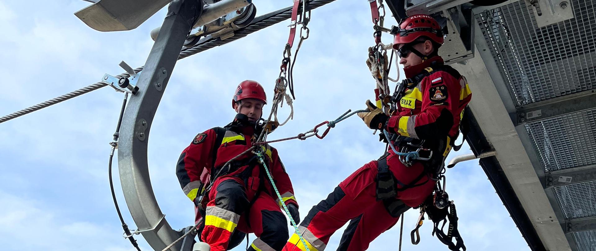 Zdjęcie przedstawia zbliżenie na dwóch ratowników wysokościowych działających na obiekcie wyciągu gondolowego. Funkcjonariusze posiadają czerwono-czarne ubranie ochronne z żółtymi elementami odblaskowymi, a ponadto hełmy oraz uprzęże, za pomocą których są przymocowani do lin wyciągu. Ratownicy na zdjęciu stoją na dachu gondoli, nad nimi można dostrzec element konstrukcji wyciągu. W tle zdjęcia widoczne są wzgórza i lasy.