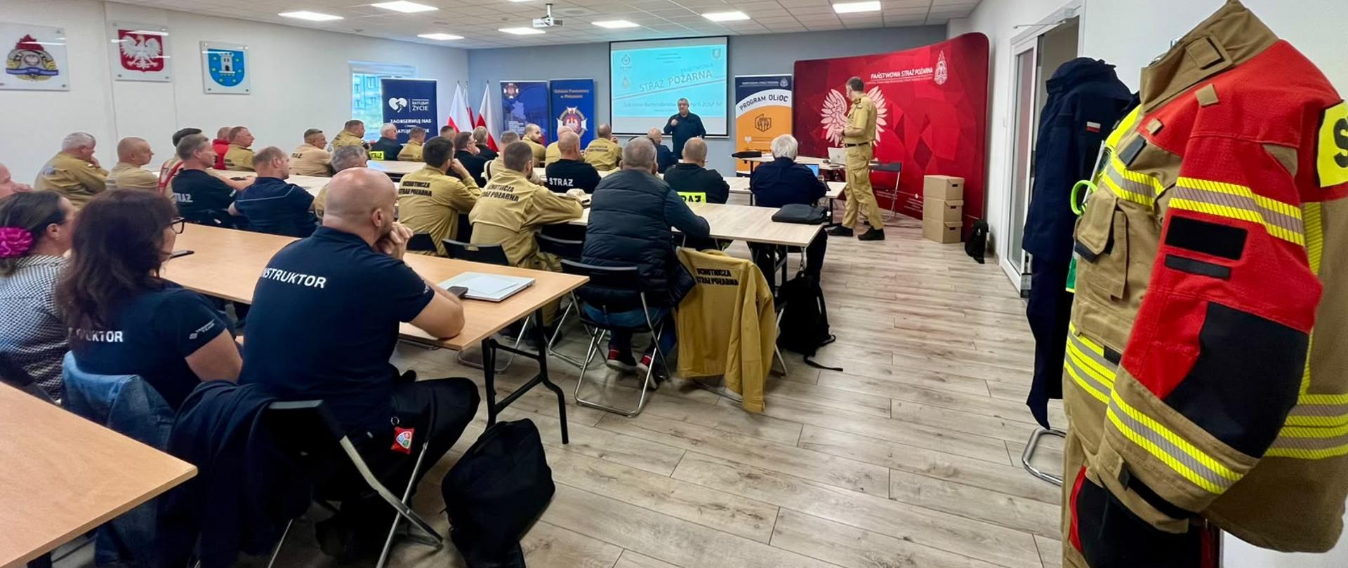 Widok z tyłu sali wykładowej na liczną grupę strażaków i instruktorów siedzących przy stołach przodem do ekranu z prezentacją „STRAŻ POŻARNA”. Na końcu pomieszczenia prelegenci prowadzą szkolenie, a na pierwszym planie po prawej stronie widać wiszący na wieszaku specjalistyczny strój strażacki.