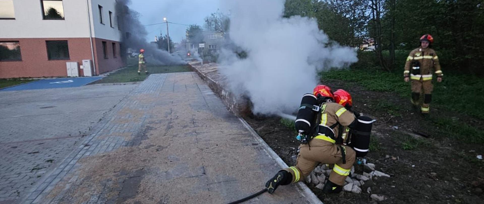 Strażacy gaszą pożar, widać dym. Z lewej strony budynek mieszkalny.