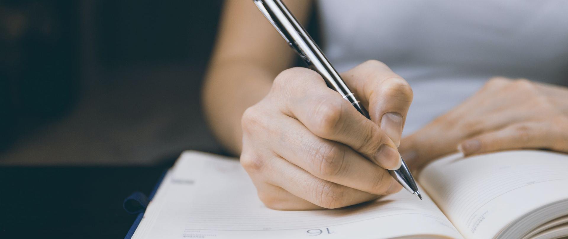 Kobieca dłoń trzymająca długopis zapisuje informacje w kalendarzu książkowym