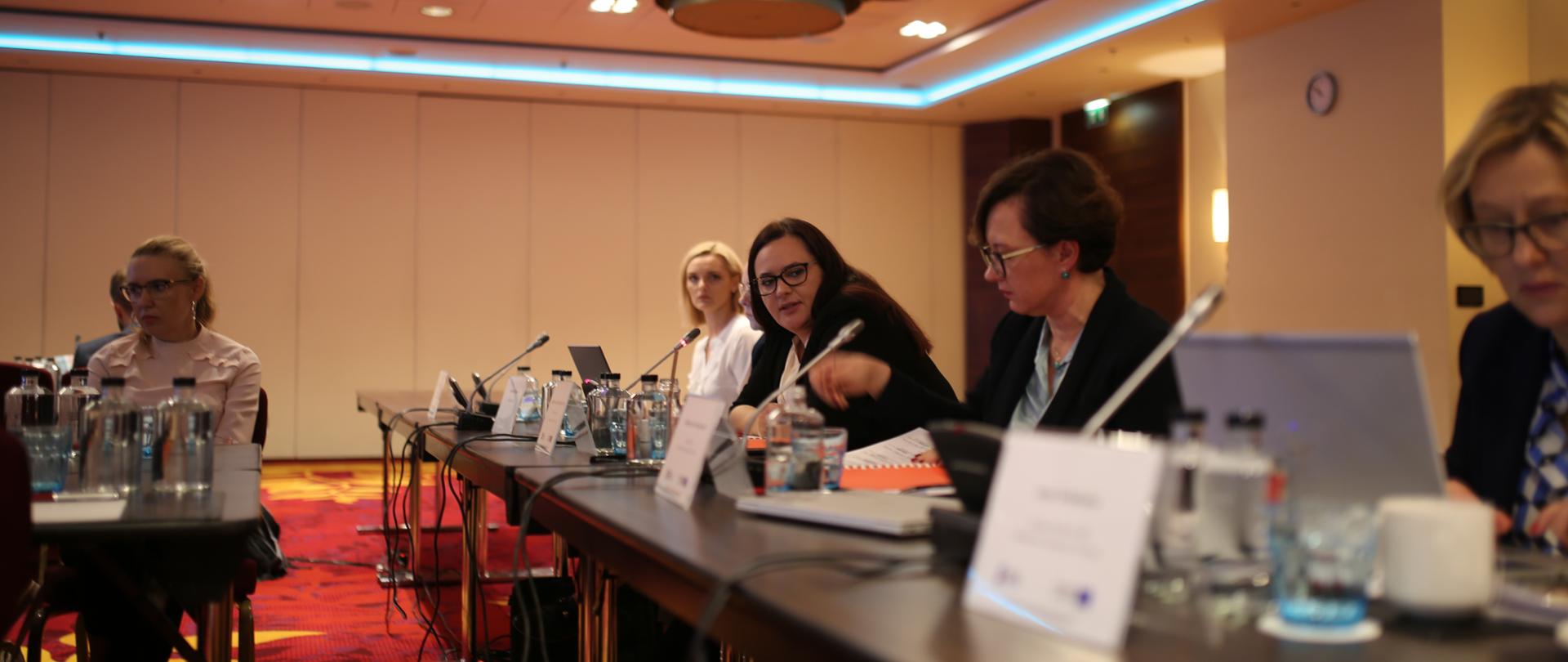 Wnętrze sali w hotelu Marriott, na podłodze czerwono-żółty dywan, na górze widoczne ledowe listwy. Na pierwszym planie długi stół, przy którym siedzi kilka osób, trzecia od prawej siedzi i mówi przez mikrofon wiceminister Jarosińska-Jedynak.