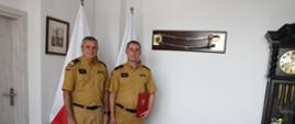 Strażacy w mundurach musztardowych stoją obok siebie jeden z nich trzyma czerwoną teczkę za nimi stoją dwie flagi Polski na ścianie wiszą obraz oraz szabla niedaleko stoi zegar.